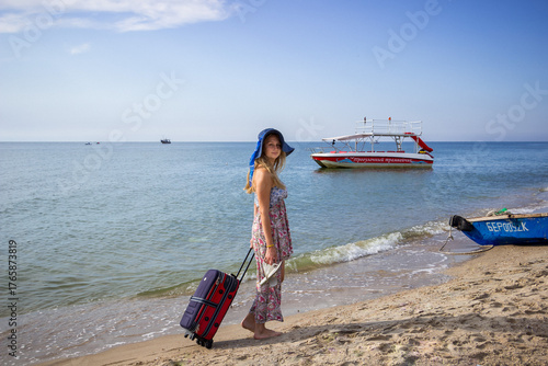 teenage girl walking on the beach, barefoot, blonde, looking back, looking into the frame, smiling, big hat on head