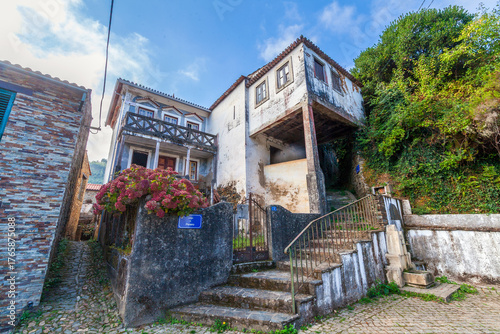 A charming, weathered stone building with a wooden balcony and vibrant pink flowers stands on a cobblestone street