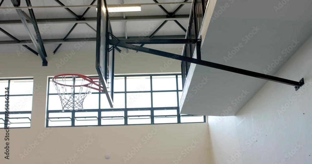 Fototapeta premium Displaying basketball hoop assembly on steel arm inside gymnasium, with exposed trusses and windows