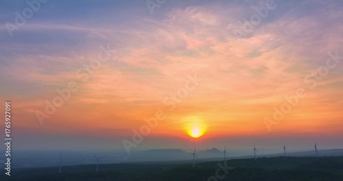 Stunning aerial scenery of sunrise above Lam Takhong Dam in Nakhon Ratchasima. where the horizon glows in soft hues. Wind turbines stand gracefully over green hills, blending nature and technology 