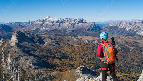 Girl with helmet and backpack admires the view from a mountain top with autumn colors in the Dolomites