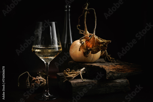 White wine glass with bottle pumpkin and dried leaves on rustic wooden blocks in moody fall setting