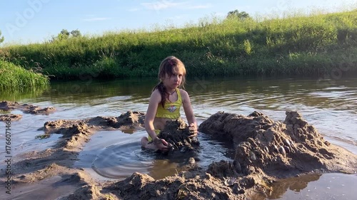 A girl in a yellow two-piece swimsuit plays in the sand on the river.