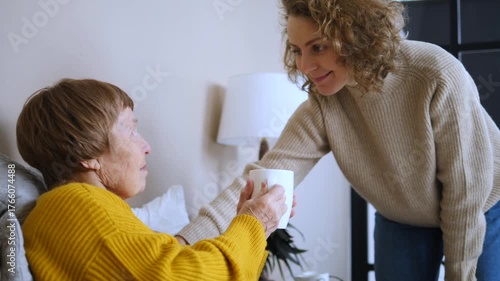 Compassionate young daughter taking care of her sick elderly mother in bed, giving her a warm cup of tea and talking, showing love, support, and family care during illness