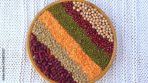 Colorful raw legumes like beans, lentils, chickpeas, and mung beans arranged in decorative stripes on a rotating wooden plate, showcasing a variety of healthy vegan protein sources