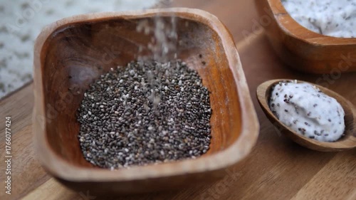 Close up of raw chia seeds being poured into a rustic wooden bowl on a cutting board, with a spoonful of prepared chia pudding visible in the background, highlighting a healthy superfood ingredient