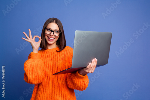 Obraz na płótnie Young woman in an orange sweater gesturing okay while holding a laptop, expressi