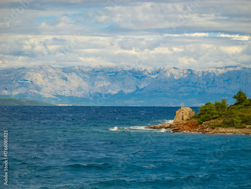 Bild auf Leinwand Old stone chapel rests on the edge of rocky shore with towering mountains in the distance, surrounded by rolling clouds