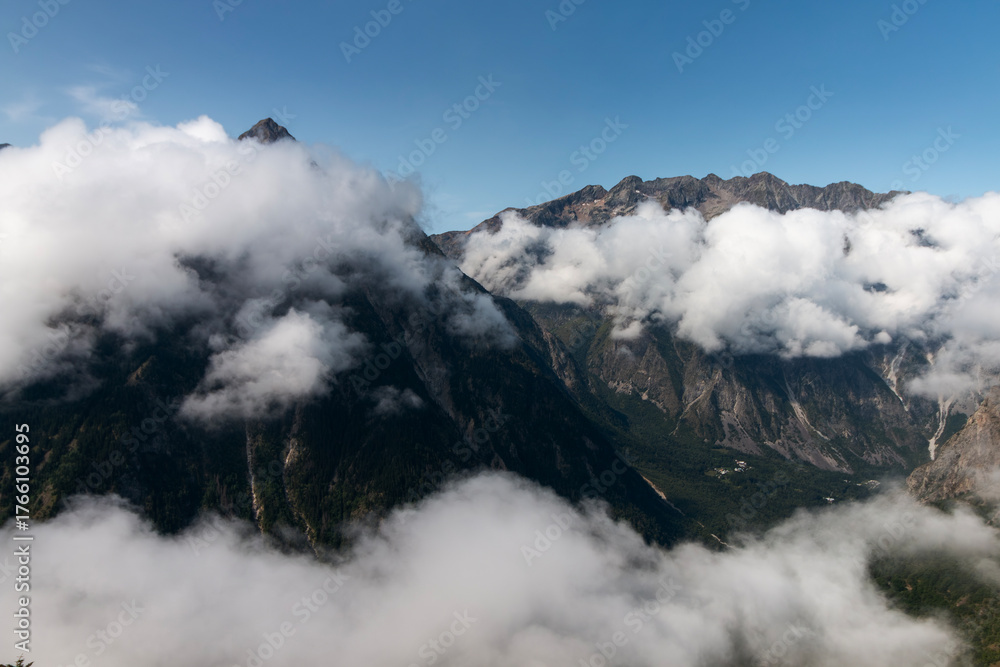 Fototapeta premium Majestic mountain peaks pierce through the scattered clouds under a clear blue sky, revealing a lush green valley below.