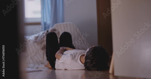 Child curled up on floor beside bed with arms resting on chest, looking tired and bored, expressing disconnection, restlessness, and lack of interest in quiet moment