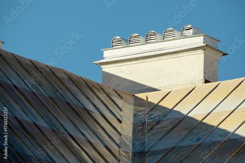 Close-up of a weathered metal roof with a ventilation chimney against a clear blue sky, showing geometric lines and sunlight shadows.