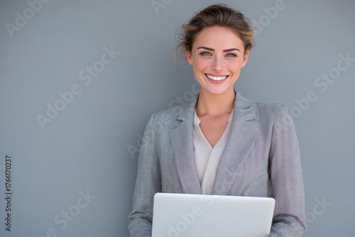 Smiling businesswoman in blazer holding laptop against neutral gray background