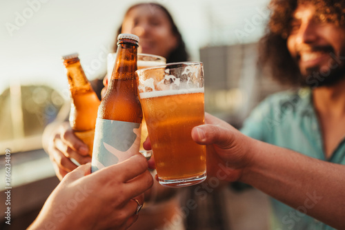 Close-up of friends clinking beer bottles and glasses during golden hour on a rooftop. 