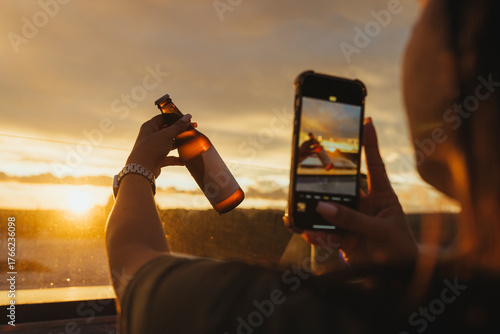 A person holds a cold beer bottle against the sunset, while another hand captures the moment on a phone. The golden light creates a warm and relaxed mood.