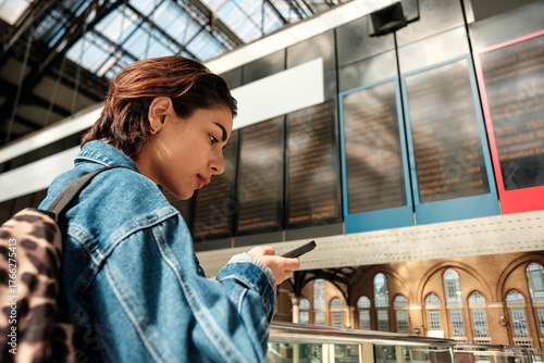 Canvas-taulu Young woman checking train schedule on smartphone in busy station