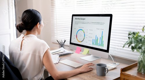 Young Asian businesswoman viewing a monthly budget report and financial charts on a desktop computer monitor in a modern office.
