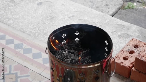 Metal urn filled with burning joss paper slowly smolders near a temple entrance surrounded by ash and smoke symbolizing offerings to ancestors. Spiritual ritual.