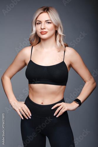Beautiful young blonde woman in black sportswear on a gray background