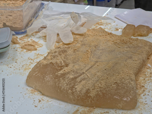 A large, translucent piece of gelatinous mochi or Warabi Mochi dusted heavily with Kinako (toasted soybean flour) on a messy preparation counter with plastic gloves.