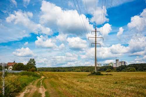 Power pylon and lines traverse a sun-kissed summer field. A winding dirt path leads towards distant city buildings under a vast blue sky with white clouds
