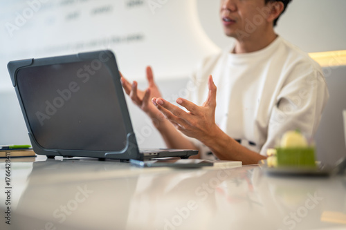 Asian man in a white shirt gesturing with both hands while speaking during an online meeting or discussion at a café table, with a laptop open in front of him in a bright workspace