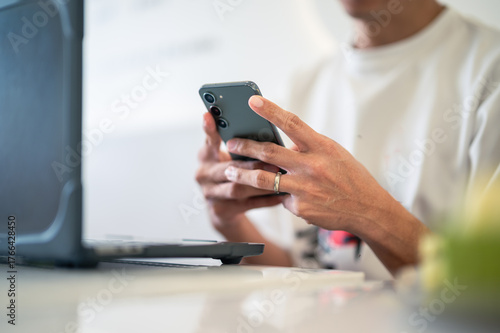 Close-up of an Asian man using a smartphone with both hands while sitting at a café table, a laptop in front of him, wearing a white shirt and a ring, showing focus in a bright workspace