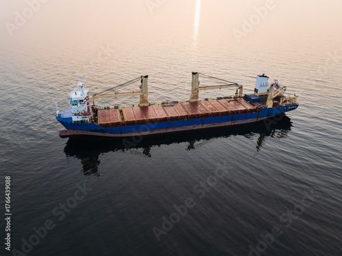 Aerial drone view of a geared bulk carrier, a dry cargo ship with cranes, sailing through the calm Baltic Sea at sunset.
