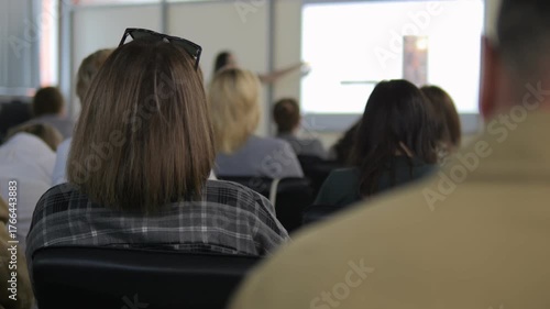 Group of people are sitting in a classroom. A woman with a plaid shirt is sitting in the front row