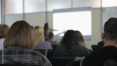 Group of people are sitting in a classroom and watching a presentation. There are several people in the room, including a man in a black shirt