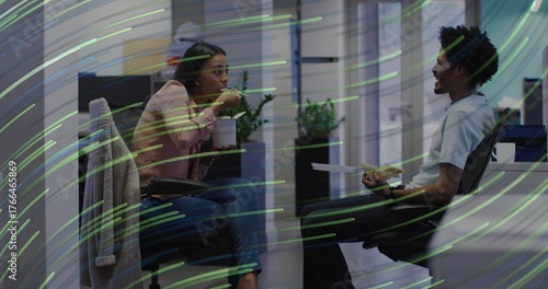 Two mid adult coworkers eating lunch in open-plan office area, with chopsticks and sandwich