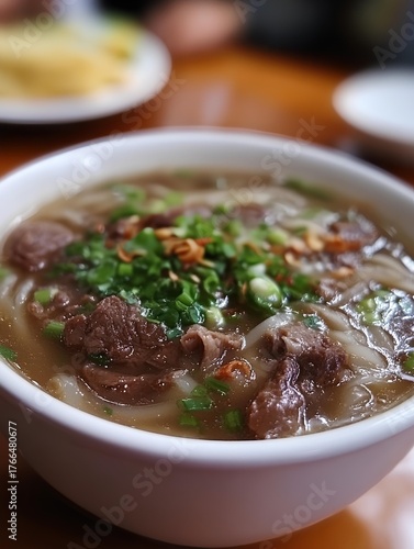 A close up of a bowl of pho with beef and green onions on a wooden table in a restaurant setting