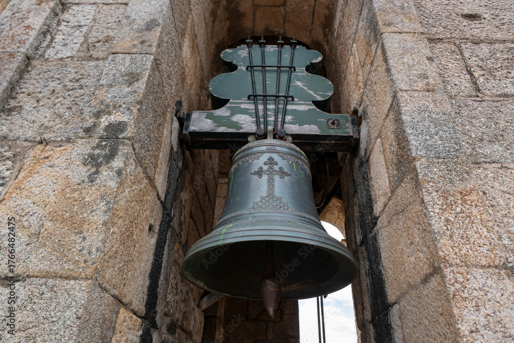 Naklejka premium Sino de bronze antigo com uma cruz a meio, suspenso numa estrutura de madeira envelhecida entre paredes de cantaria rústica numa torre de uma Igreja