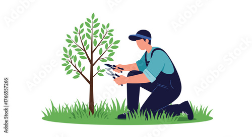 A man wearing a cap and overalls is kneeling in green grass, carefully pruning the branches of a young tree with pruning shears in a garden.