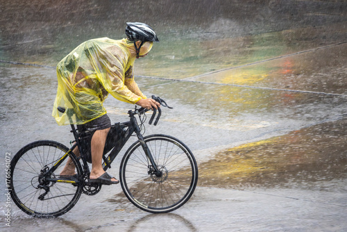 Fototapeta Naklejka Na Ścianę i Meble -  Cyclist in a raincoat rides through heavy rain on a city street