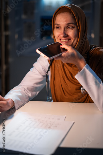 Concept of Artificial intelligence in medicine. Muslim doctor in hijab command to ai assistant, dictate audio message using smartphone working on night shift at clinic