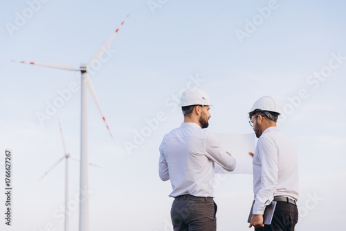 Engineers discussing blueprint in front of wind turbines, exploring sustainable energy solutions