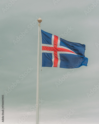 View of the Icelandic flag ripples against a pale sky, its blue and red cross a stark contrast to the muted tones, Arnarstapi, Snaefellsbaer, Iceland.