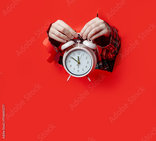 Merry Christmas. classic white alarm clock is held in hands wearing plaid sleeves, emerging from torn red paper background.