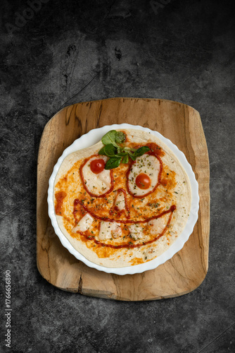 Close-up of Halloween-themed pizza with smiling face made from vegetables and cheese on white plate over wooden board, top view food photography