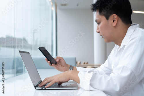 Focused man working with laptop and smartphone in modern office, representing multitasking, digital business, online communication, remote work, mobile technology, and professional productivity.