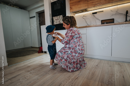 Bilde på lerret A mother kneels in a floral dress, sharing a drink with her child in a sunlit, modern kitchen