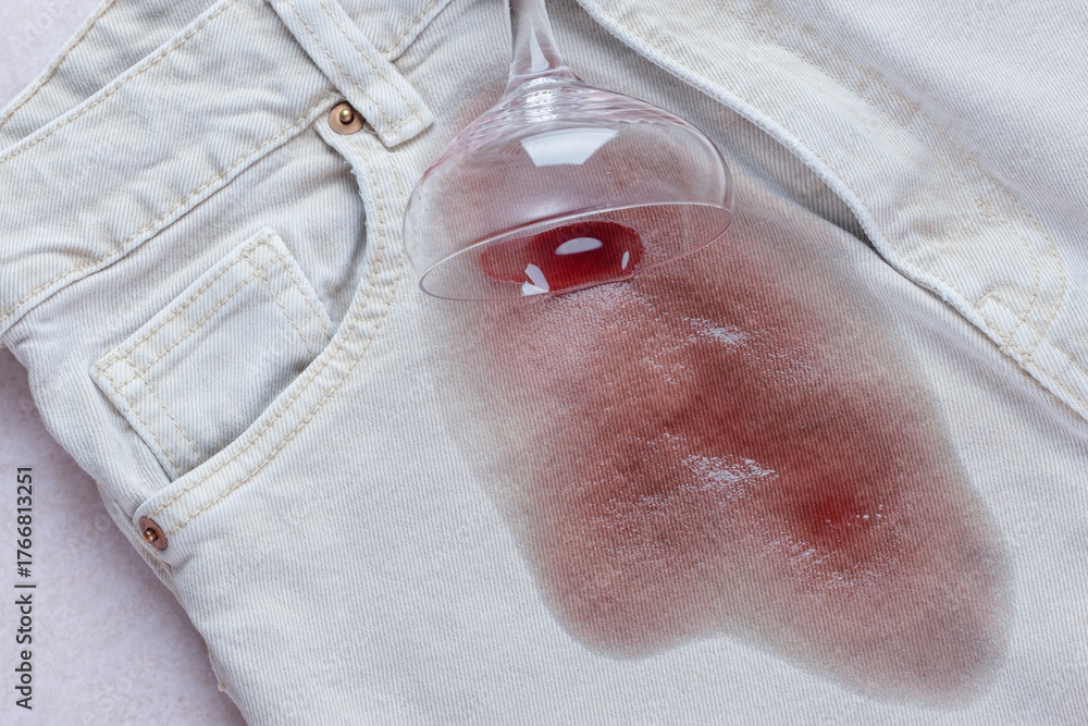 Fototapeta premium Spilled glass with red wine falled a top a light blue pair of jeans, while a noticeable red stain spreads from the glass. Daily stains concept. top view