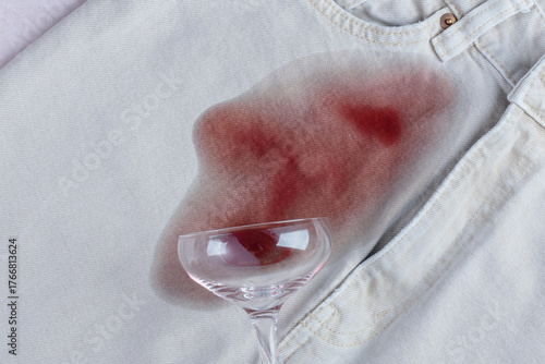 Spilled glass with red wine falled a top a light blue pair of jeans, while a noticeable red stain spreads from the glass. Daily stains concept. top view