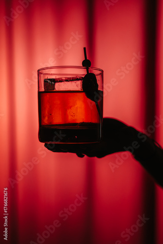 Photos Close-up of a classic cocktail with ice and garnish in a glass, held against a red background