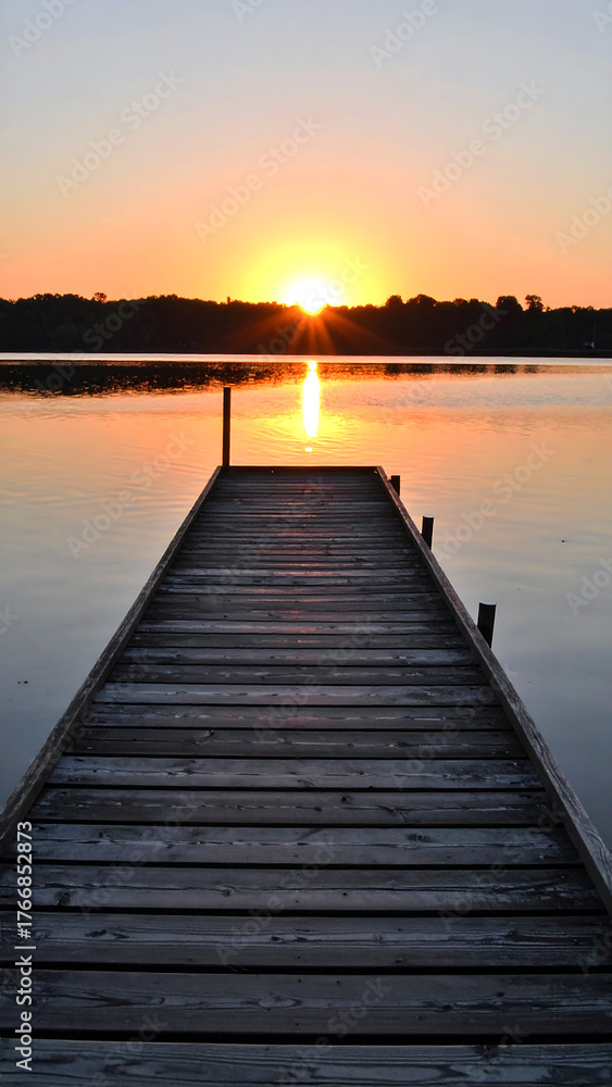 Fototapeta premium Wooden pier extending into calm water during sunrise with orange sky