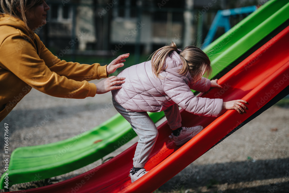 Fototapeta premium A caring mother helps her little daughter climb up a vibrant slide at a park. This joyful moment captures family bonding and the excitement of exploring playground adventures.