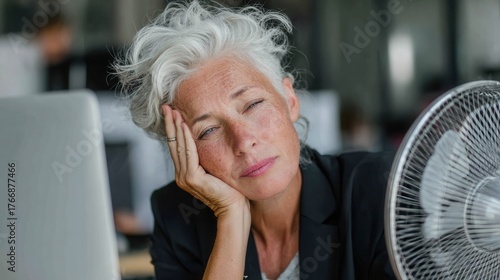 Mature businesswoman experiencing menopause symptoms at work, hot flush and sweating in modern office, health awareness, stress, hormone balance, fatigue, resilience