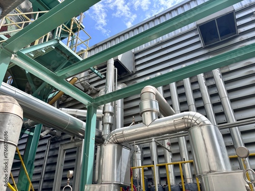 Insulated stainless steel industrial piping system with elbows and valves, featuring green structural steel beams against a metal factory wall and blue sky. This complex network transfers fluids 