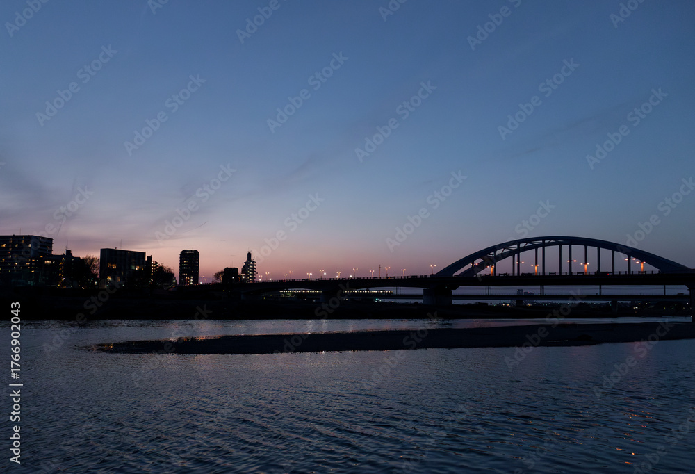 Fototapeta premium City bridge at twilight over calm river, evening lights and skyline silhouette, Japan/夕暮れに照らされた都市の橋と静かな川の風景