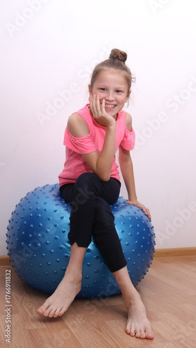 A little girl sitting on a blue balance ball during home workout. Child practicing balance and coordination, developing motor skills and posture.